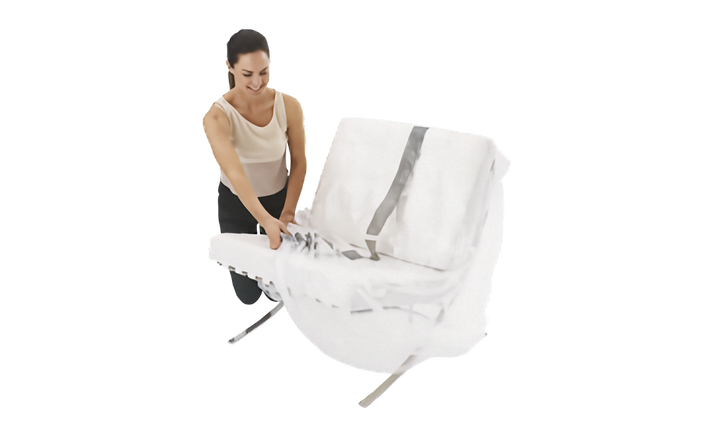 A woman kneels beside a white upholstered chair with a white seat cushion and metal legs. The woman is pulling a clear lounge chair cover with grey middle stripe over the chair, shown against a plain white background.
