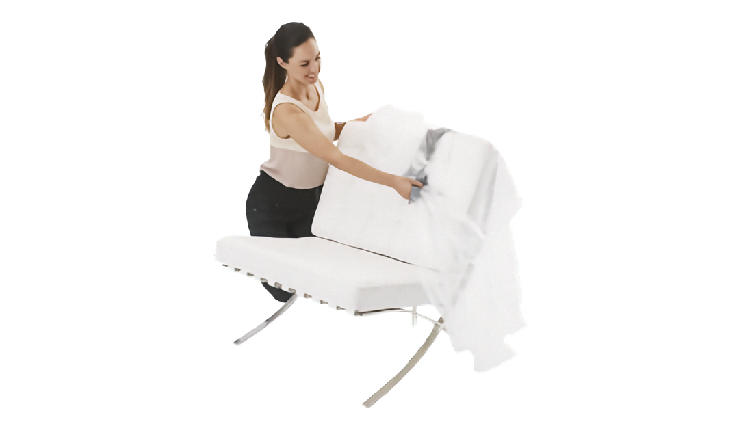 A woman in a beige tank top and black skirt is shown behind a white upholstered chair place a clear plastic lounge chair cover over the chair. The background is a clean, solid white studio setting.