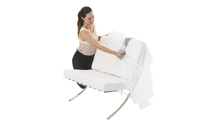 A woman in a beige tank top and black skirt is shown behind a white upholstered chair place a clear plastic lounge chair cover over the chair. The background is a clean, solid white studio setting.