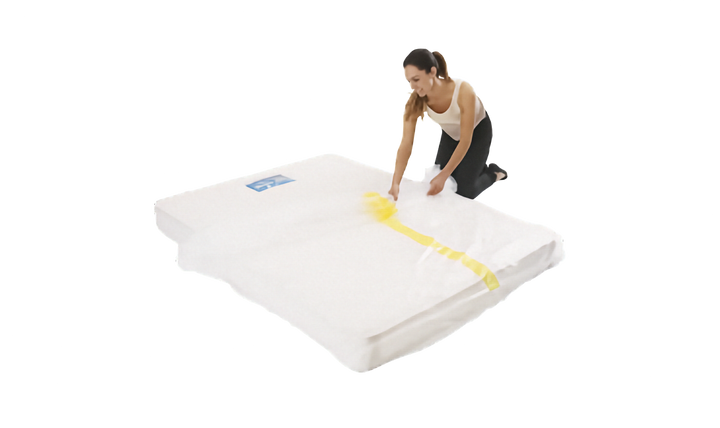 A person is shown sliding a clear plastic mattress protector onto a mattress. The cover has a yellow strip. Shown on a plain white background.