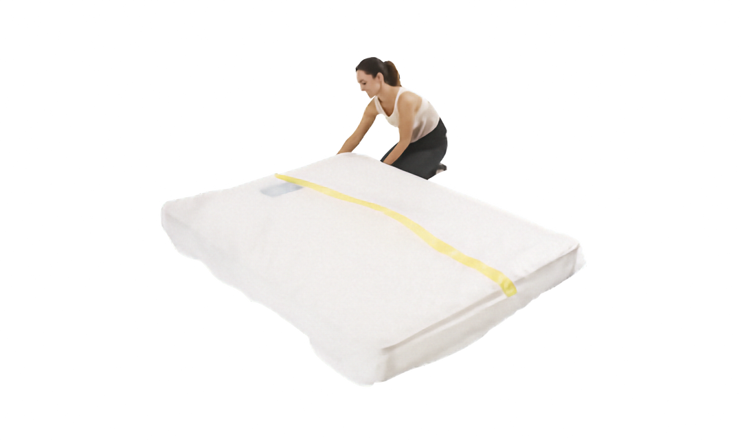 A person is shown sliding a clear plastic mattress protector onto a mattress. The cover has a yellow strip. Shown on a plain white background.